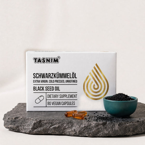 Tasnim Schwarzkümmelöl Kapseln, Packung mit 80 veganen Kapseln auf Basaltplatte, davor Kapseln und Petrol-Schale mit Schwarzkümmelsamen