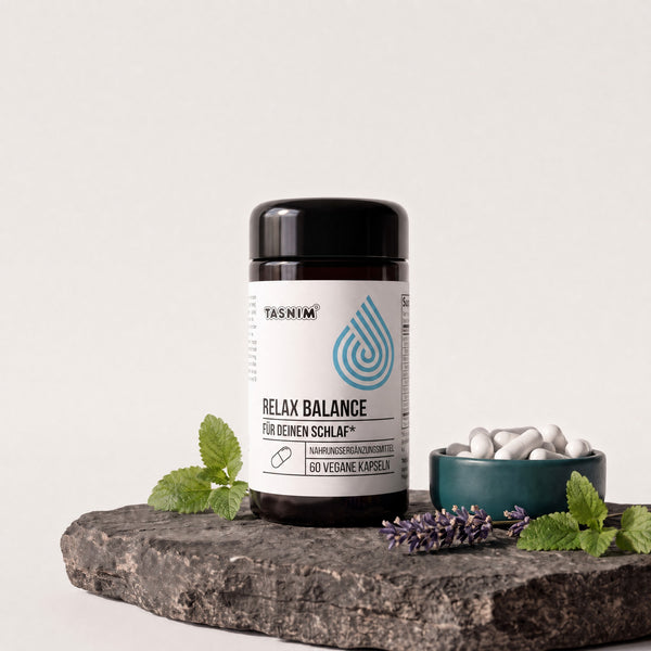 Tasnim Relax Balance, Dose mit 60 veganen Kapseln auf Basaltplatte, daneben eine Petrol-Schale mit Kapseln, Lavendel und Zitronenmelisse