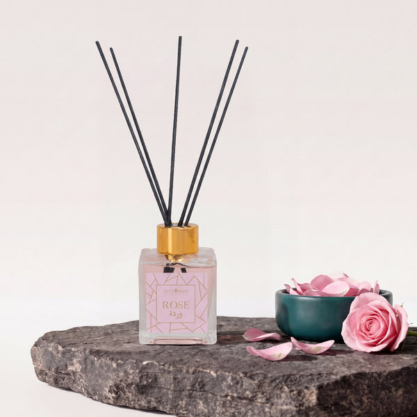 Duftstäbchen Rose: Glasflakon mit Goldkappe, schwarzen Rattanstäbchen, rosa Flüssigkeit und Rose-Etikett auf dunkler Steinplatte, daneben Rosenblätter