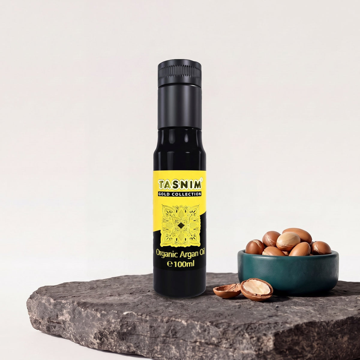 Tasnim Arganöl Gold Collection, 100-ml-Glasflasche mit gelb-schwarzem Etikett auf Basaltplatte, daneben eine Petrol-Schale mit Argannüssen