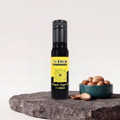Tasnim Arganöl Gold Collection, 100-ml-Glasflasche mit gelb-schwarzem Etikett auf Basaltplatte, daneben eine Petrol-Schale mit Argannüssen