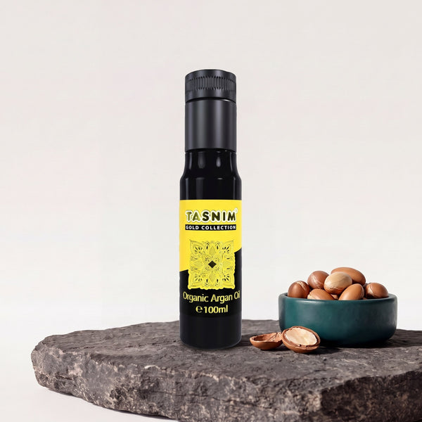 Tasnim Arganöl Gold Collection, 100-ml-Glasflasche mit gelb-schwarzem Etikett auf Basaltplatte, daneben eine Petrol-Schale mit Argannüssen