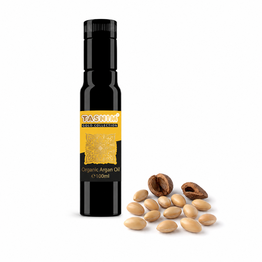 Schwarze 100-ml-Flasche Tasnim Bio Arganöl mit goldenem Gold-Collection-Etikett neben losen Argankernen und zwei aufgebrochenen Arganschalen auf weißem Hintergrund.
