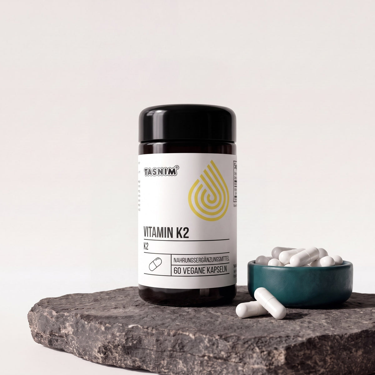 Tasnim Vitamin K2, Dose mit 60 veganen Kapseln auf Basaltplatte, daneben eine Petrol-Schale mit weißen Kapseln