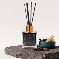 Duftstäbchen Black Oud: Glasflakon mit Goldkappe, schwarzen Rattanstäbchen, dunkler Flüssigkeit auf dunkler Steinplatte, daneben Oud-Holz und Sandelholz