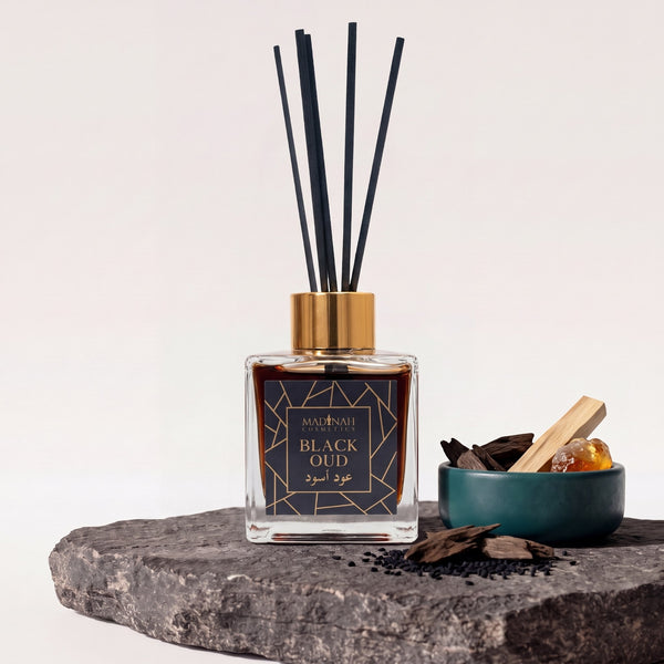 Duftstäbchen Black Oud: Glasflakon mit Goldkappe, schwarzen Rattanstäbchen, dunkler Flüssigkeit auf dunkler Steinplatte, daneben Oud-Holz und Sandelholz